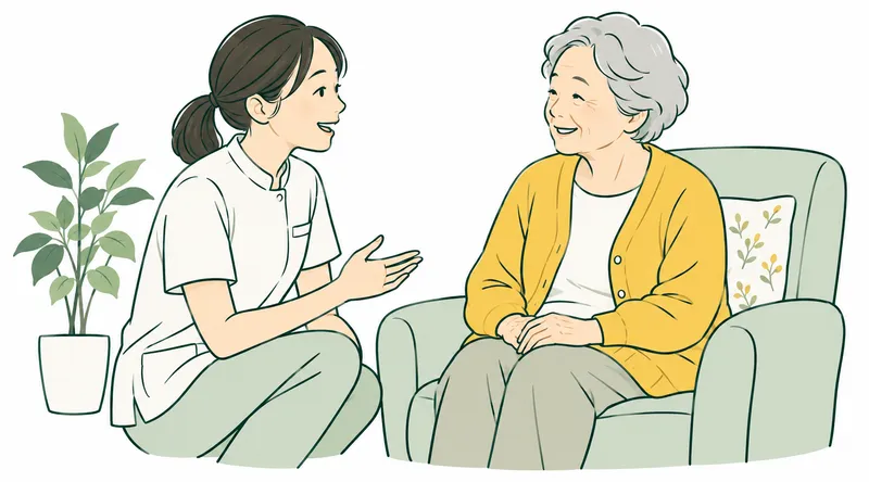 A smiling Japanese care professional in a simple work tunic talking at eye level with an elderly person seated in an armchair. Warm, equal, dignified interaction.